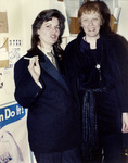 Leighty with a tie, tuxedo, and cigarette in-hand poses with Reeves in a long, velvet jacket and scarf around her waist, both smiling. Posters and displays are in the background.
