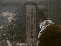 A person kneels at a wooden grave marker with black calligraphy, in front of a mound of dirt and straw.