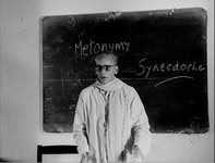 In black and white cinematography, a teacher stands in front of a classroom chalkboard with "metonymy" and "synecdoche" written on it in white English. The chalkboard's edges are covered in half-way erased marks leftover from previous writings.