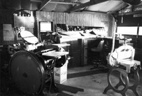 Black-and-white view of the print shop: letterpress, paper cutter, type cabinets, ink cabinet on the wall, trim saw. Small window and row of windows along the garage door.