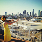 Color image of a woman in a yellow dress playing a viola. She is on a roof and overlooks the skyline of Los Angeles. She faces away from the viewer.