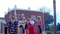 Four women dressed in a variety of vibrant colors dance outdoors in front of a dark red and white building.