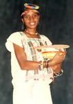 Young woman wearing Fulani woman’s handwoven dress. The dress is white, embroidered with diamond patterns between vertical bands of black, green, and red thread.