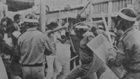 Three helmeted policemen with shields and batons are seen from the back charging at demonstrators who appear startled, angry, and hopeless.