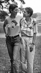 Black-and-white outdoor photo. The two are standing, arms around each other, looking toward each other and smiling. Both are in short-sleeved shirts and long pants.