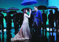 A color photo of a stage performance. It depicts two figures holding an umbrella together at the center and a group of people holding umbrellas in the background.