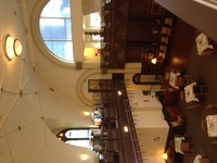 Photo taken from dark wood-railed balcony, overlooking tables set for four. Walls and vaulted ceiling are ivory-colored.