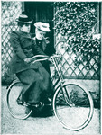 Postcard depicts Willard with her new bicycle, Gladys. A friend assists as she pedals, dressed in a long skirt and bonnet. Background shows lattice with ivy.