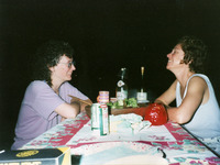 Color photo. The two sit in the dark at a picnic table facing each other with big smiles on a summer evening. Salad bowl, candles, and beverages cover a print tablecloth.