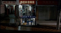 A street front at night, with individuals visible inside the shops. Calligraphy is visible on the signs and posters.