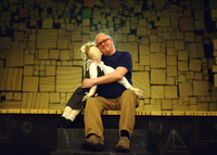 Man in a black T-shirt sitting on a theatrical stage smiling with a life-sized fabric doll.