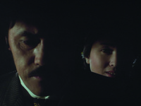 Another very low key composition revealing, in extreme close-­up, half of Aochi’s countenance, facing the camera with downcast eyes, Screen Left. The other half vanishes in shadow. Right, Sono’s countenance, lit by a single strong beam, appears behind Aochi’s shoulder with a questioning, sly expression.