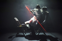 Figure 16.2. Lit from one side, a man sits on a folding chair with a woman in a white dress on his lap. Behind them, the robot KUKA shines a red laser beam cutting a diagonal across them.