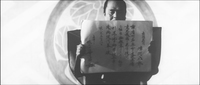A samurai reads from an unrolled proclamation. He stands before a large family crest.