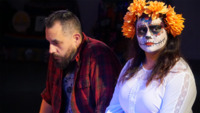 A man in a flannel shirt sits and looks down. Beside him is a young woman with her face painted as a Day of the Dead skeleton and with a marigold wreath in her hair.