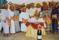 A group of women wearing aso ebi headscarves and shawls gather around a table with cakes on it.
