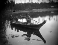 A black-and-white photograph of a family of three in a dugout canoe: two adults and one small child. One of the adults is throwing a double pronged sealing spear into the water.