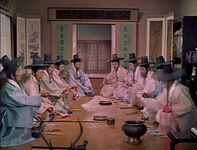 A group of men sit before screens, a couplet, and a framed work of calligraphy, shot in near-perfect symmetry.