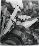 A black-and-white photograph of a figure using a crooked knife to carve a piece of wood.