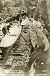 A black-and-white photograph of two men working to make dugout canoes. Each man is working on a separate canoe and two other canoes are in the background to the side.