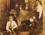 Early twentieth century era sepia-toned photograph, eight men gathered around a scenic cut-out of a cow, one underneath pretending to drink from the cow’s udder.