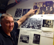 Man in a black shirt pointing to a sepia-toned, old photograph posted on a wall of a group of men in three-piece suits
