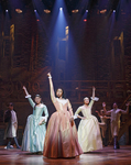 The three Schuyler sisters in taffeta dresses. Actor Phillipa Soo wears light blue, Renée Elise Goldberry wears peach, and Jasmine Cephas Jones is in light yellow. Each sister has one hand in the air, making the gesture “Work it.”