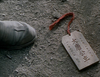 A wooden tag with red string and black calligraphy on the ground, at a person's foot.