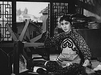 Woman sitting in the bed of a Shaanxi cave house. Her right hand turns a cotton spinning wheel and her left hand holds a piece of cotton that feeds the wheel.