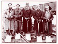 Five Black women dressed in warm street clothes, gloves, and various head coverings stand on railroad tracks, all with shovels. They look at the viewer. Train cars in the background.