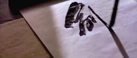 A film still of calligraphic text being written on a sheet of paper, with the brush visibly on a downstroke.
