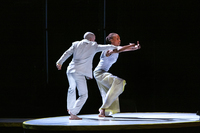 A moment in Chamberlin and Jenkins’s duet on the circular satellite stage, brightly lit while the rest is in darkness. Jenkins’s arms move as though wrapping around a large basket; she bends at the waist as she brings her right knee upward, looking over her hands into the audience. Chamberlin stands with his back to the audience and reaches his right arm into the basket shape to disrupt Jenkins’s gesture; his left hand is in a fist.