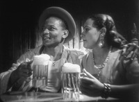 A black and white film still features a multiracial man and woman seated at the Siete Mares Club in Konga roja. They are both holding full mugs of beer and the man in the hat has his arm around the woman.