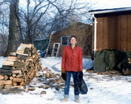 Color photo. Cohen stands, smiling, behind the house in a winter scene with snow. Next to her are two or three cords of wood that she has split for the wood-burning stove.