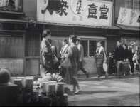 Calligraphic signs over a public street, on which families and other passersby are strolling. In the foreground there is a stack of cans.