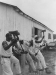 Black-and-white photograph of a group of incarcerated African American men with raised axes, singing in the woodyard of a prison farm