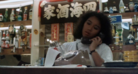 A shopgirl in a medium shot holds the receiver of a rotary phone to her ear, with white calligraphy on a black banner right above her head.