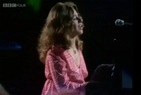 Figure 13. Carole King, wearing a pink dress, sits at the piano and sings with her eyes closed and an expression of deep immersion in the music on her face.