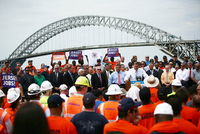 Christie next to a podium surrounded by workers wearing hard hats and signs saying “Jersey Jobs!”