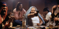 Medium shot of Kallu mama (Saurabh Shukla) firing a gun to the right of the camera in “Goli Maar Bheje Mein,” in Satya. He is surrounded by fellow gang members as they smoke, drink, and gamble.