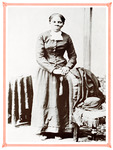 Studio portrait. Tubman stands, hands resting on a chair, in a long skirt, a long-sleeved buttoned jacket, and scarf with her hair braided and pulled back. Intense gaze toward the viewer.