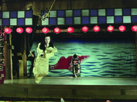 Complex long shot of a theatrical mise-­en-­scène. In the background the head of the “puppet Shinako” (see Figure 8.23) hangs down, with the same ocean backdrop behind. A black clad female puppeteer manipulates a black curtain screen Right. Middle ground, a ghostly Oine in funereal white clothes and painted white face glides out from upper Left backstage on a transom, crossing at an angle towards the top Left of screen and past the viewer.