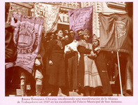 Tenayuca, arm raised in a fist, speaks on the steps of City Hall surrounded by other organizers, both women and men, some holding large satin banners for the Alliance of Workers.