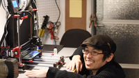 Tony Loiseleur, who identifies himself as “a product of the French occupation of Vietnam,” wears glasses, a driving cap, and a black sweater. He kneels in front of a work station with a light gray surface with a yellow stripe at the edge. On the work station, eye level with Tony, is a 3D printer with dark brown and red plastic parts and metal hardware. Behind Tony is a dark gray metal garage door. Tony rests his right forearm and left hand on the work station surface.