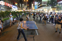 This photograph shows two individuals playing table tennis in the middle of a street at night. The street is crowded with pedestrians and several people are watching the game.