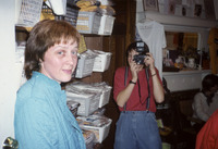 Color photo taken in the shop. Reeves looks away from the photographer to the viewer with a soft smile. Bins of T-shirts sit beside her and postcards are displayed on the mantel.