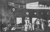 Production photograph depicting large ship set piece with portholes and actors gathered in foreground.