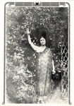 Ethereal full-length photo. Duncan stands under an arbor with thick foliage. Her floral print gown is draped over one shoulder. One arm is extended up. Her hair is pulled back.