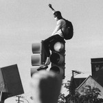 Image depicts a Black young man with fist raised sitting on top of a traffic light. Photo in black and white. The man is in focus, blurred foreground of another fist in the air, this one with lighter skin, likely a white person. Houses and trees and electric wires are visible in the background.