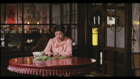 Image of a woman sitting alone at a round wooden table in a restaurant, doing prep work with a bowl of greens. Through a window in the deep background, there are pink and red banners with black calligraphy on them.